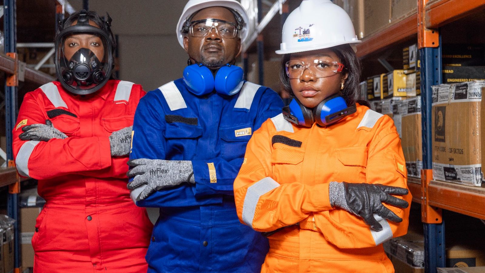 Três profissionais da RIGSTOCK com equipamento de protecção individual — fatos de trabalho, capacetes, óculos, luvas e máscara — num armazém.